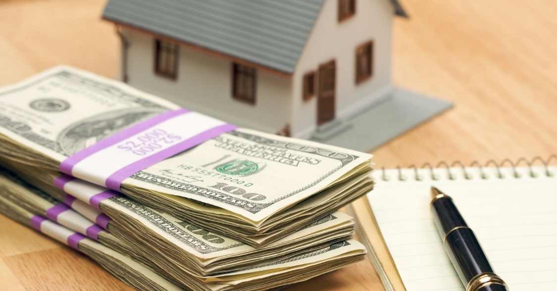 Stacks of $100 bills in front of a small model of a house